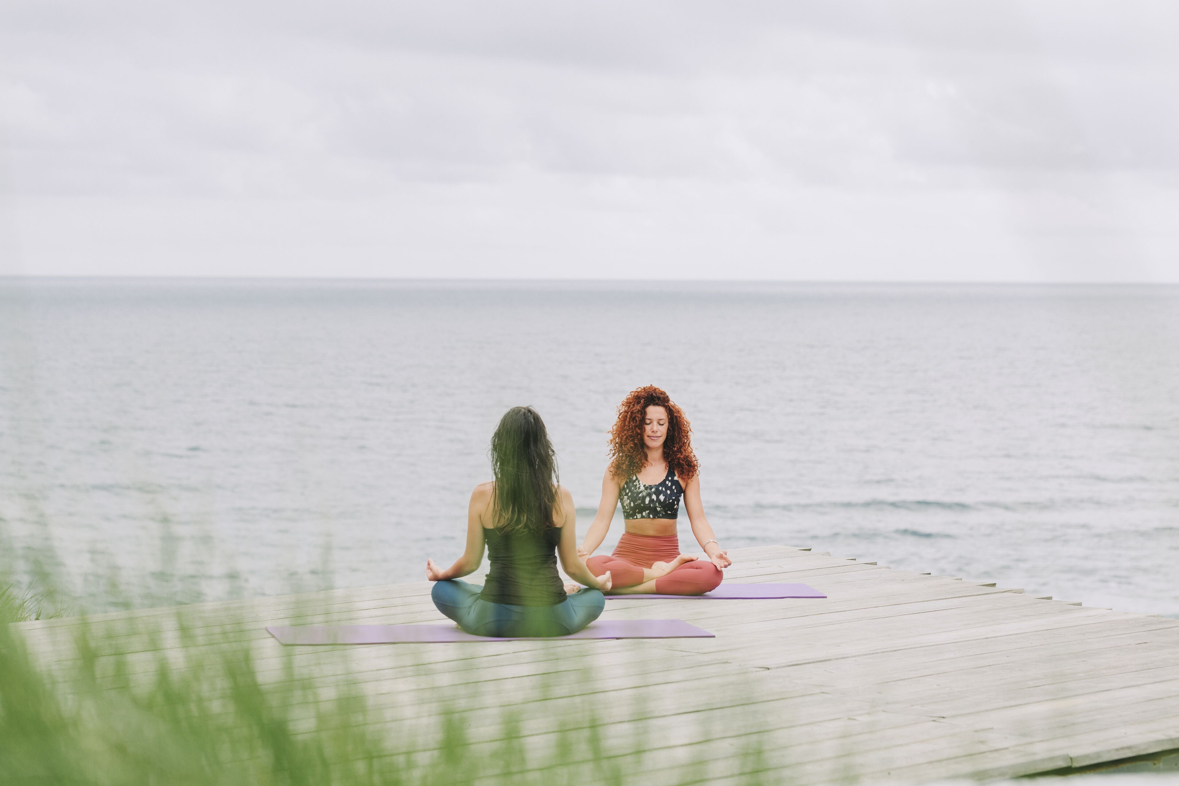 Two individuals sitting on yoga mats on a wooden dock by the sea.