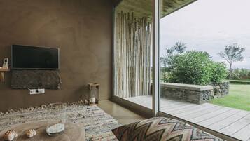 Una sala de estar moderna con un televisor grande, una mesa de vidrio con caracoles, una alfombra estampada y vistas a un jardín a través de amplios ventanales.