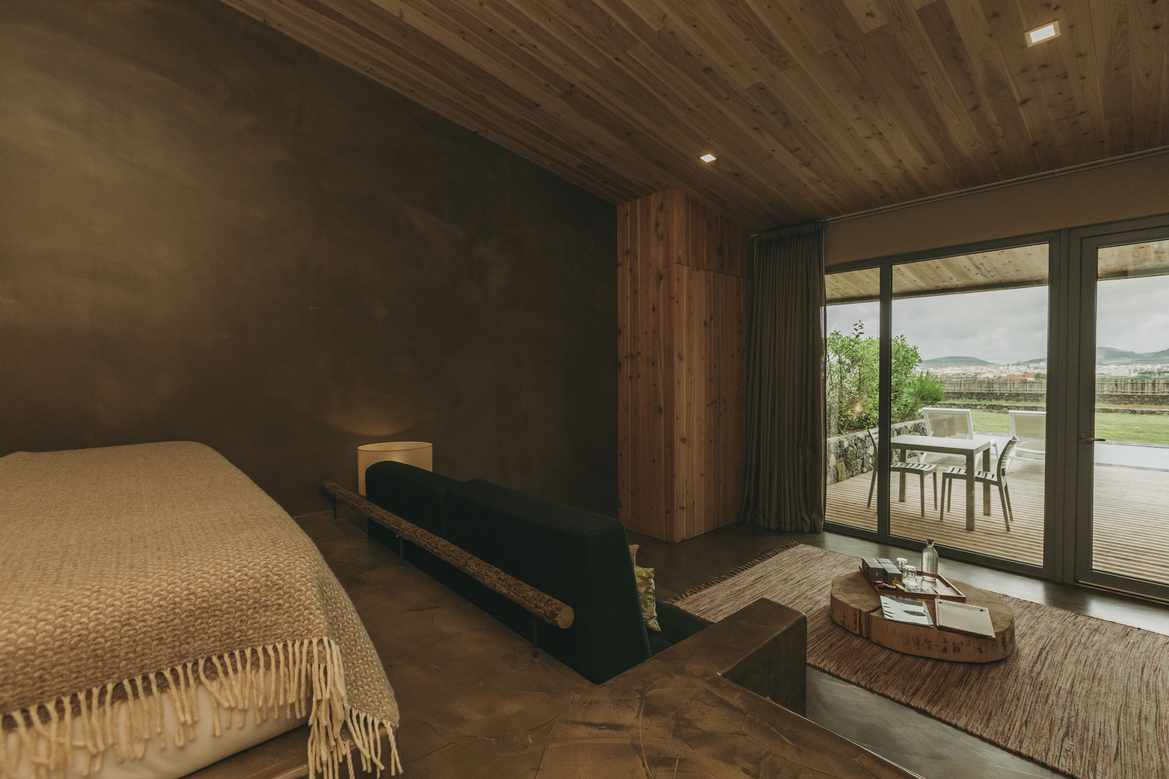 A modern bedroom with a large bed, a wooden headboard, a small seating area with a table, and a view of the outdoors through large windows.