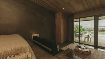 A modern bedroom with a large bed, a wooden headboard, a small seating area with a table, and a view of the outdoors through large windows.