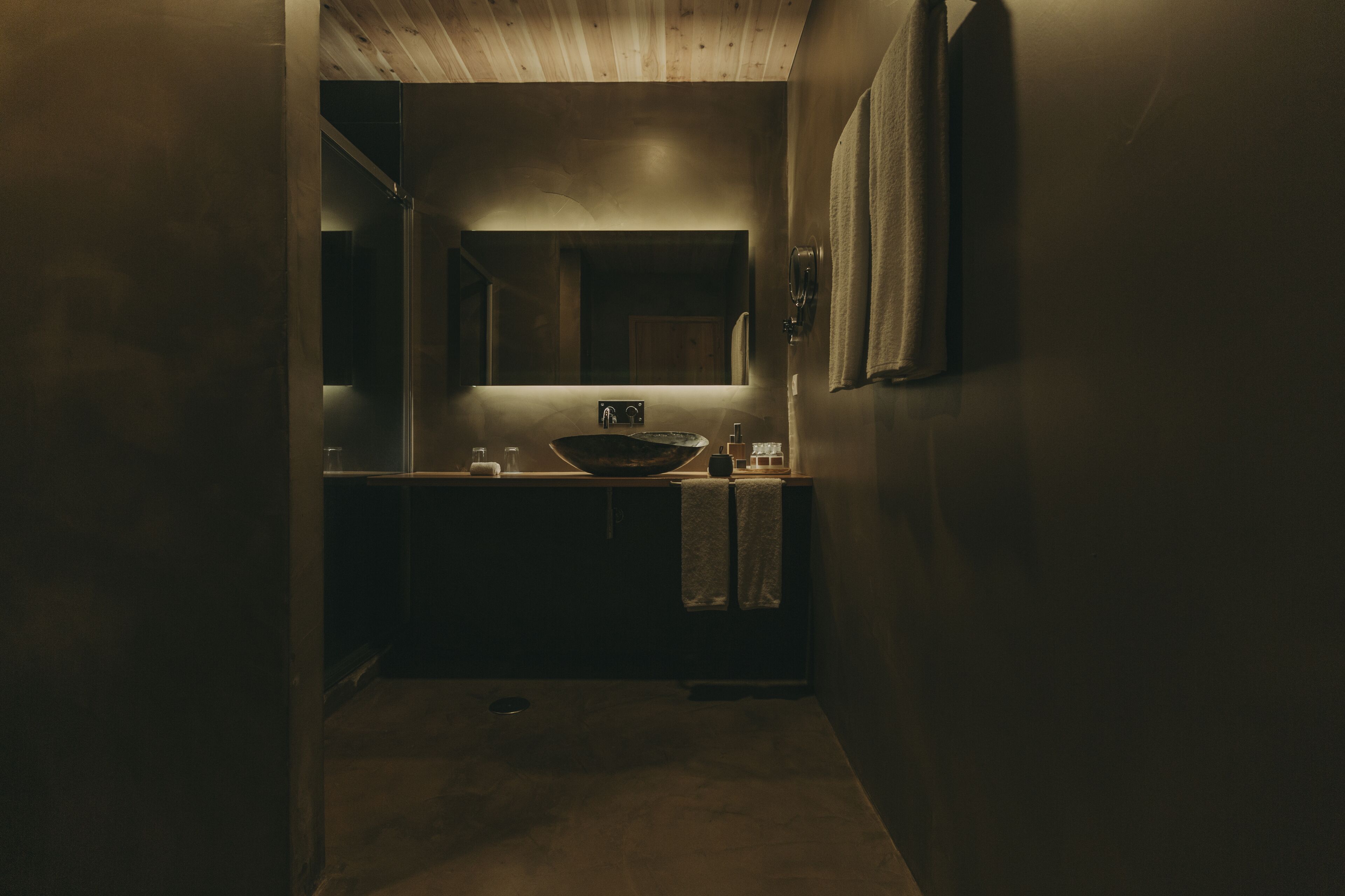 Un pasillo estrecho y tenuemente iluminado que da a un baño con un tocador de madera, un espejo grande y una toalla colgada en la pared.