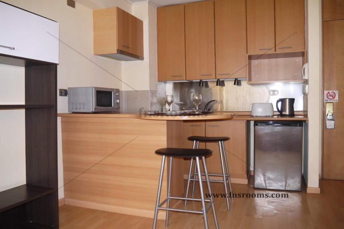 A compact kitchen with wooden cabinets, a microwave, a coffee maker, and two stools.