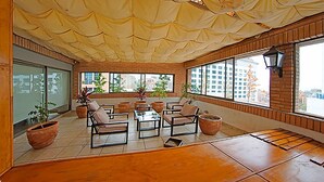 A spacious indoor area with large windows, multiple seating arrangements, and potted plants. - Trani Apart Las Condes (Santiago)