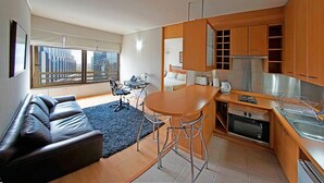 A modern kitchen with wooden cabinets, a dining area with a black couch and chairs, and a large window offering a city view. - Trani Apart Las Condes (Santiago)