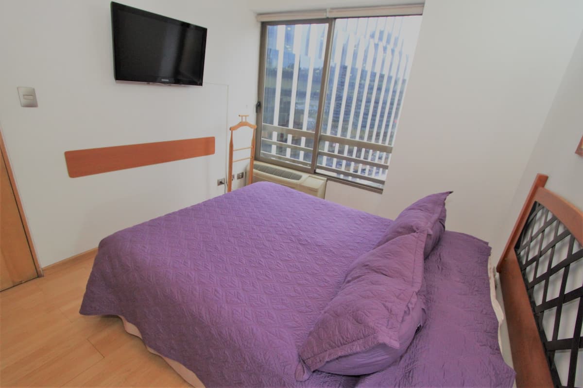 A bedroom with a bed, a TV mounted on the wall, and a window with a view of buildings.