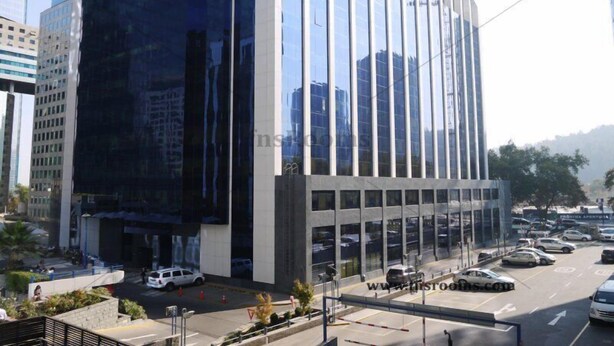 A modern building with reflective glass windows and a parking lot with several cars. - Trani Apart Las Condes (Santiago)
