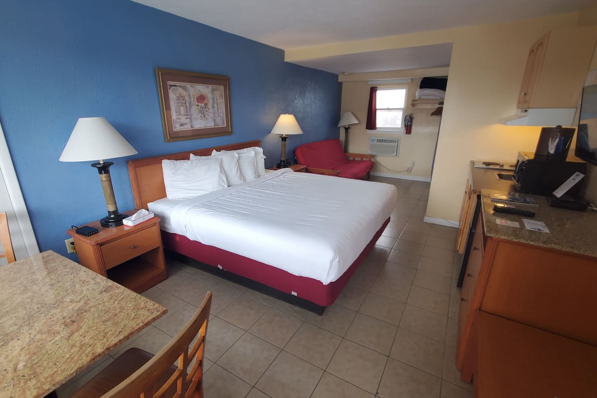 A hotel room with a bed, bedside tables, a desk, chairs, a kitchenette, and a television.