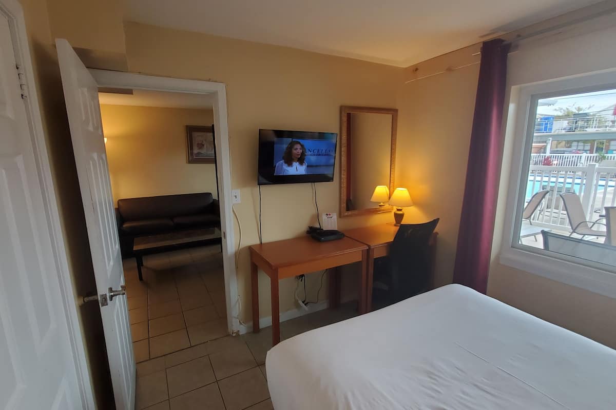 A hotel room with a bed, a desk with a TV, a chair, a lamp, and a window with a view of a pool and buildings.