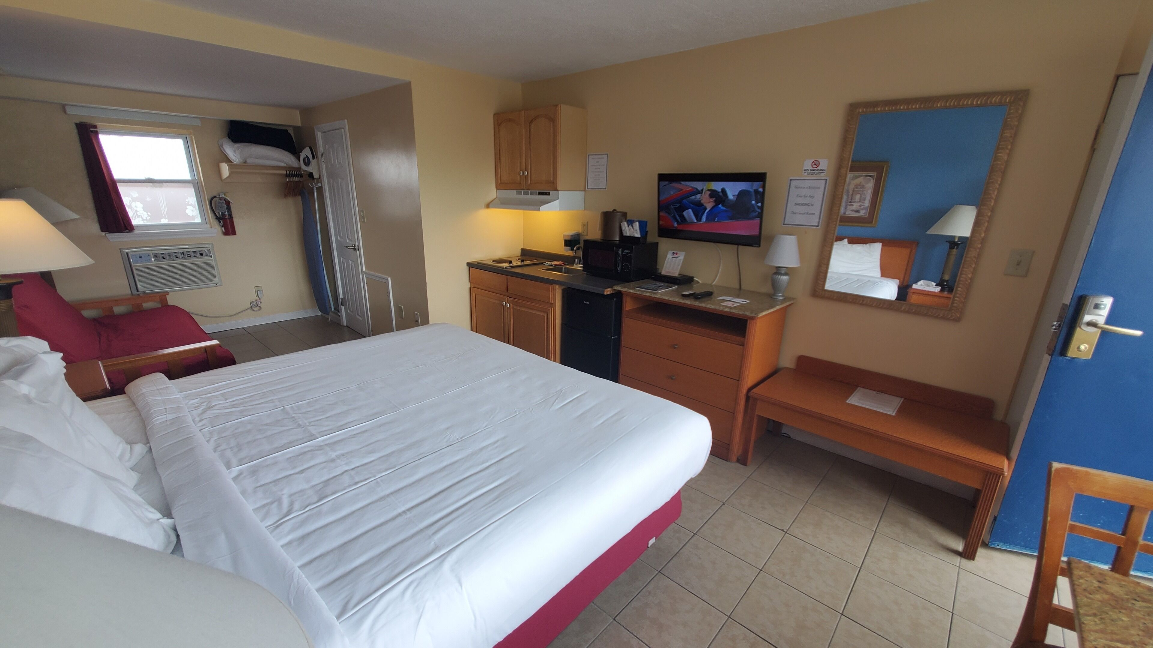 A hotel room with a bed, a desk, a chair, a television, and a kitchenette.