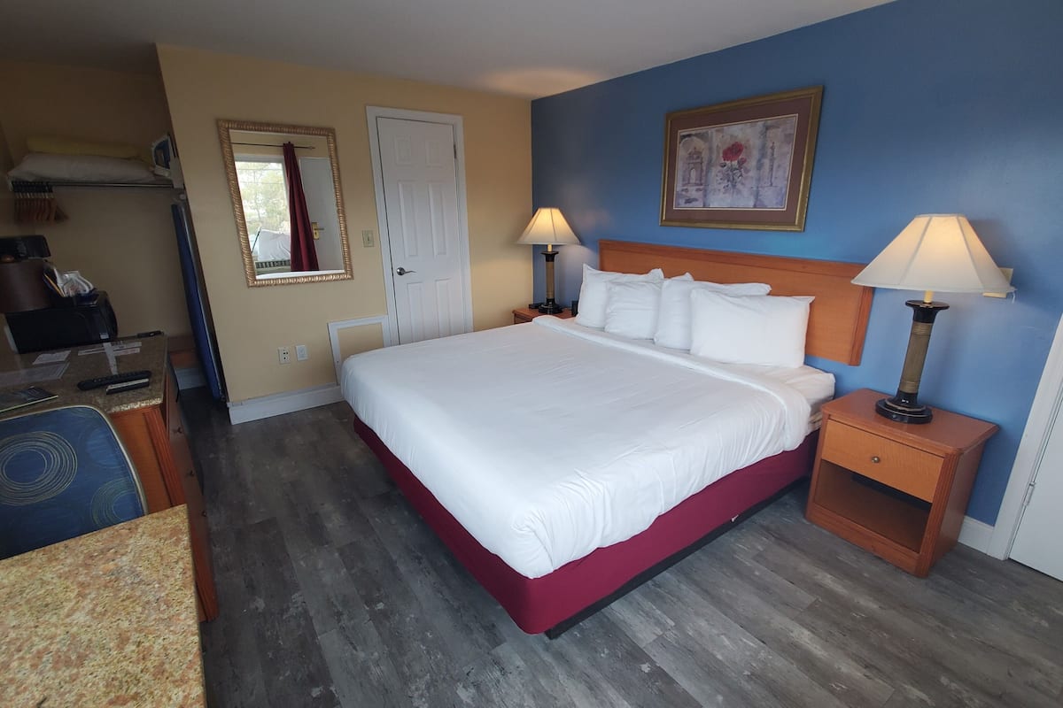 A hotel room with a large bed, two bedside lamps, a wooden nightstand, a desk with a chair, a mirror, and a door.