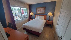 A hotel room with a bed, two bedside lamps, a nightstand, a desk, and a window with a view of the outside. - Sand Box Motel (Wildwood)