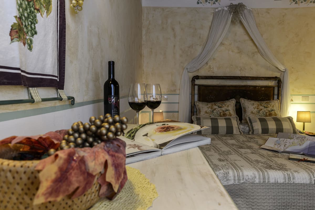 A bedroom with a bed, a bedside table with a wine bottle and glasses, a decorative basket, and a canopy bed.