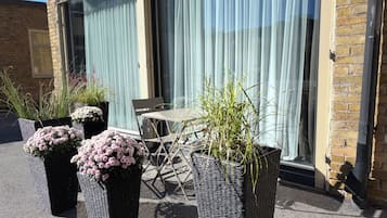 Uteplatser med blomsterprydda korgplanterare, ett bord och en stoluppsättning, placerade framför en byggnad med en fasad av tegel och stora fönster.