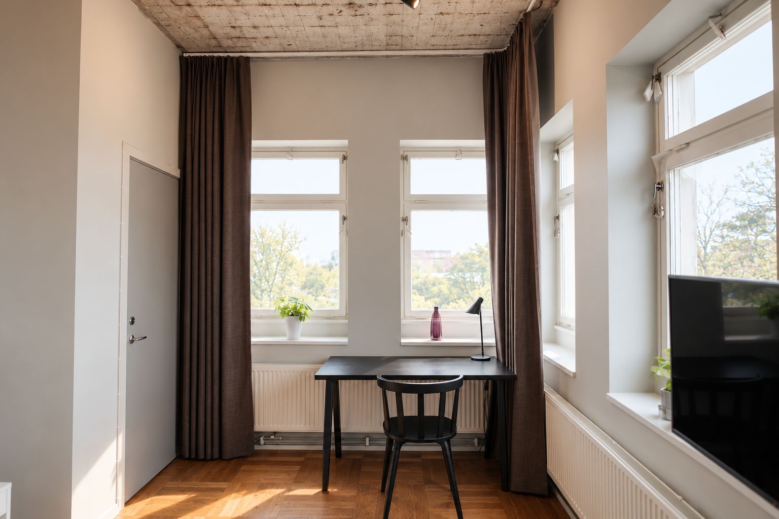 Ein Zimmer mit Holzboden, Schreibtisch mit Stuhl, zwei Fenster mit Vorhängen, Heizkörper und Fernseher.