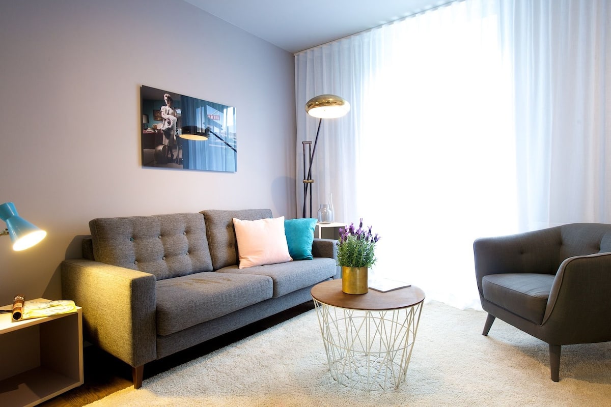 Een moderne woonkamer met een grijze bank, een ronde salontafel en een bijzettafeltje met een lamp.