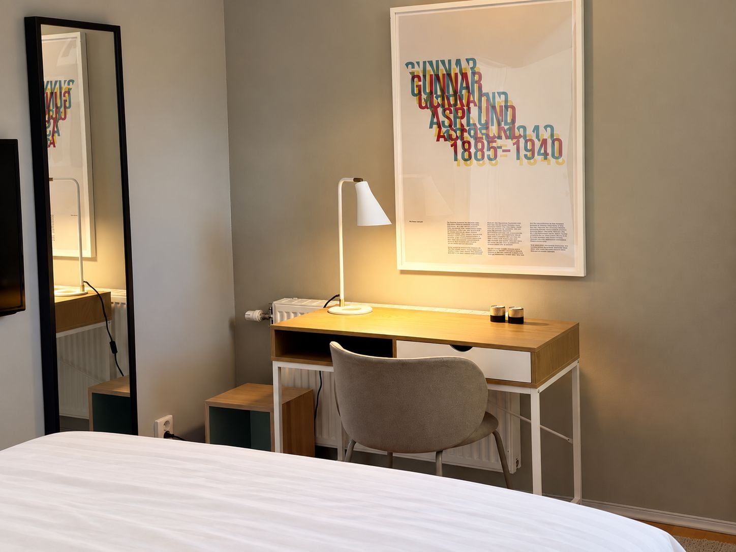 Una camera d'albergo con un letto, una scrivania con lampada, una sedia, uno specchio grande e un quadro appeso al muro.