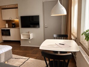 Una habitación moderna con una mesa de comedor, un televisor montado en la pared, una zona de cocina y un ventanal con cortinas.