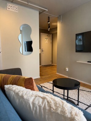 Una sala de estar moderna con un sofá, una mesa redonda de centro, un televisor montado en la pared y un pasillo que conduce a una puerta.