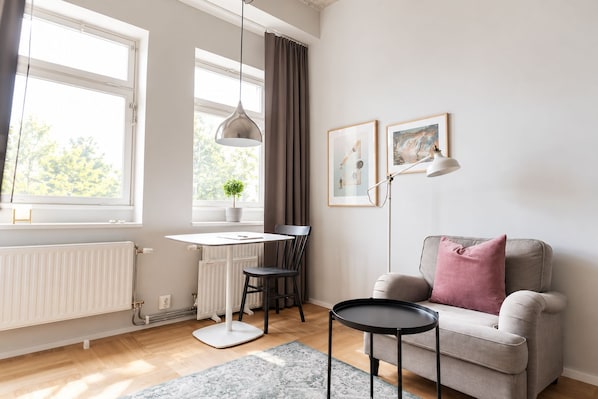 A modern living room with a white radiator, a small table, a grey sofa with a pink pillow, and two framed pictures on the wall. - Asplund Hotel Apartments (Solna)