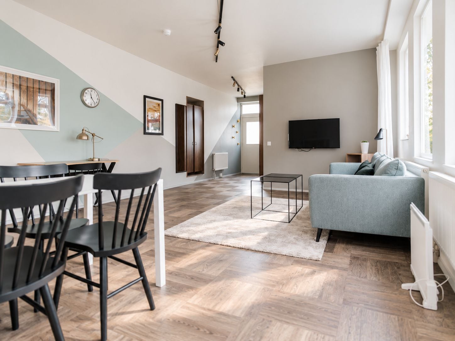 Una sala de estar moderna con un sofá gris, una zona de comedor con sillas, un televisor de pantalla plana montado en la pared y una escalera que sube a otro nivel.