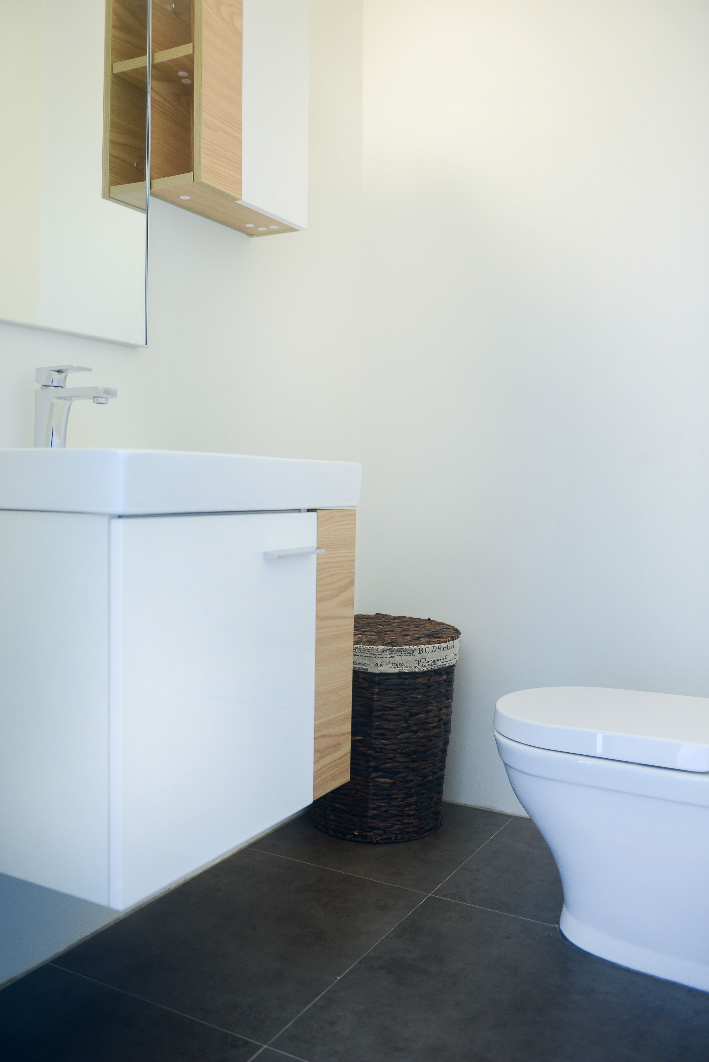Une salle de bain moderne avec un lavabo blanc, un meuble en bois, une poubelle en osier et des toilettes blanches.