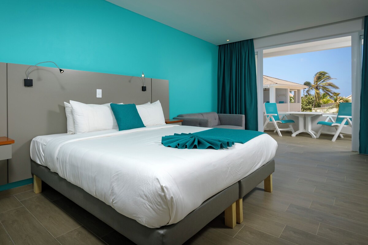 A modern hotel room with a large bed, a desk, and a balcony with outdoor seating.