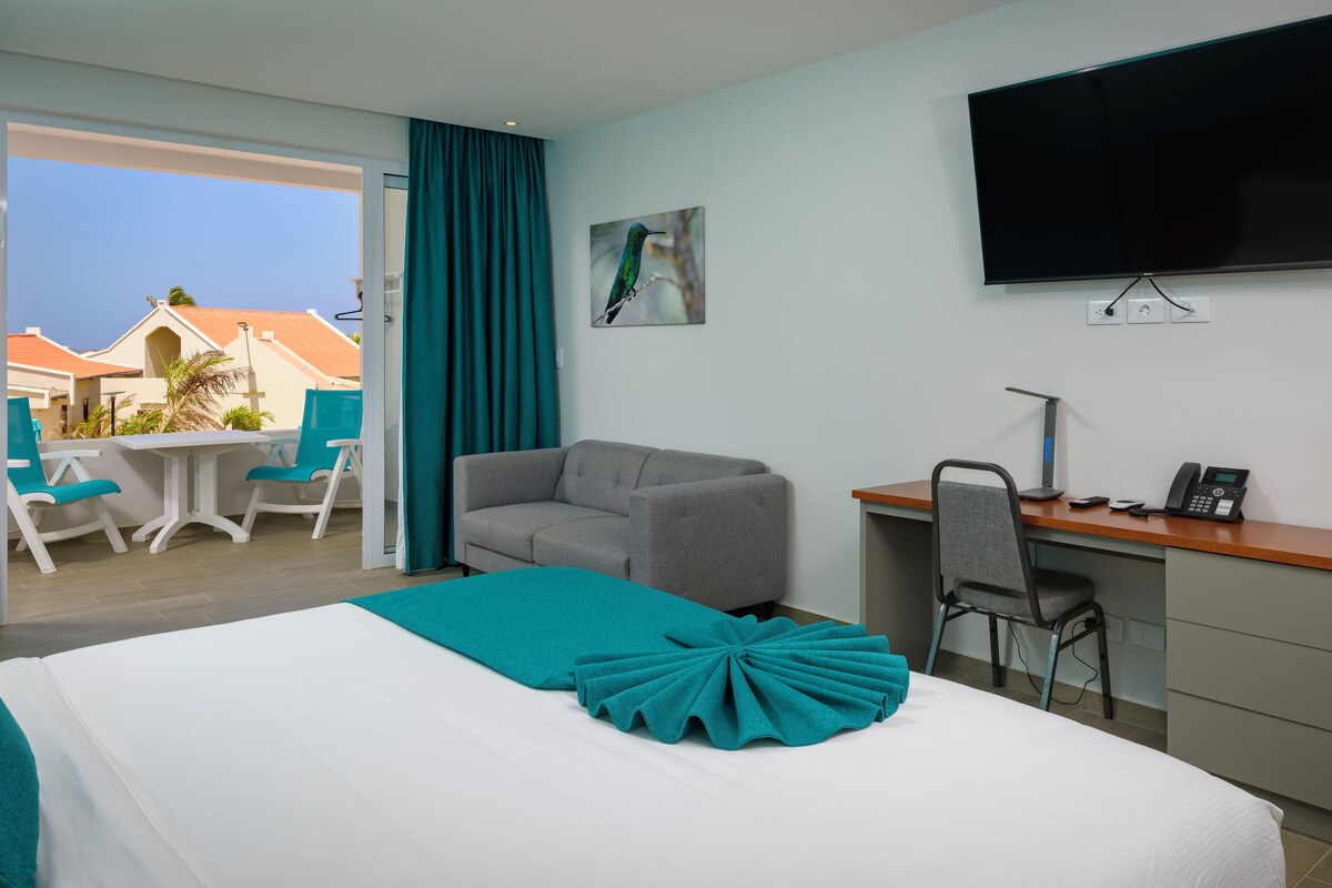 A hotel room with a bed, a desk, a chair, a TV, and a balcony with a view of buildings.