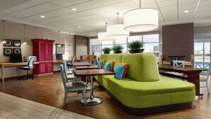 A modern lounge area with a green sofa, wooden tables, and chairs. There are potted plants and pendant lights.
