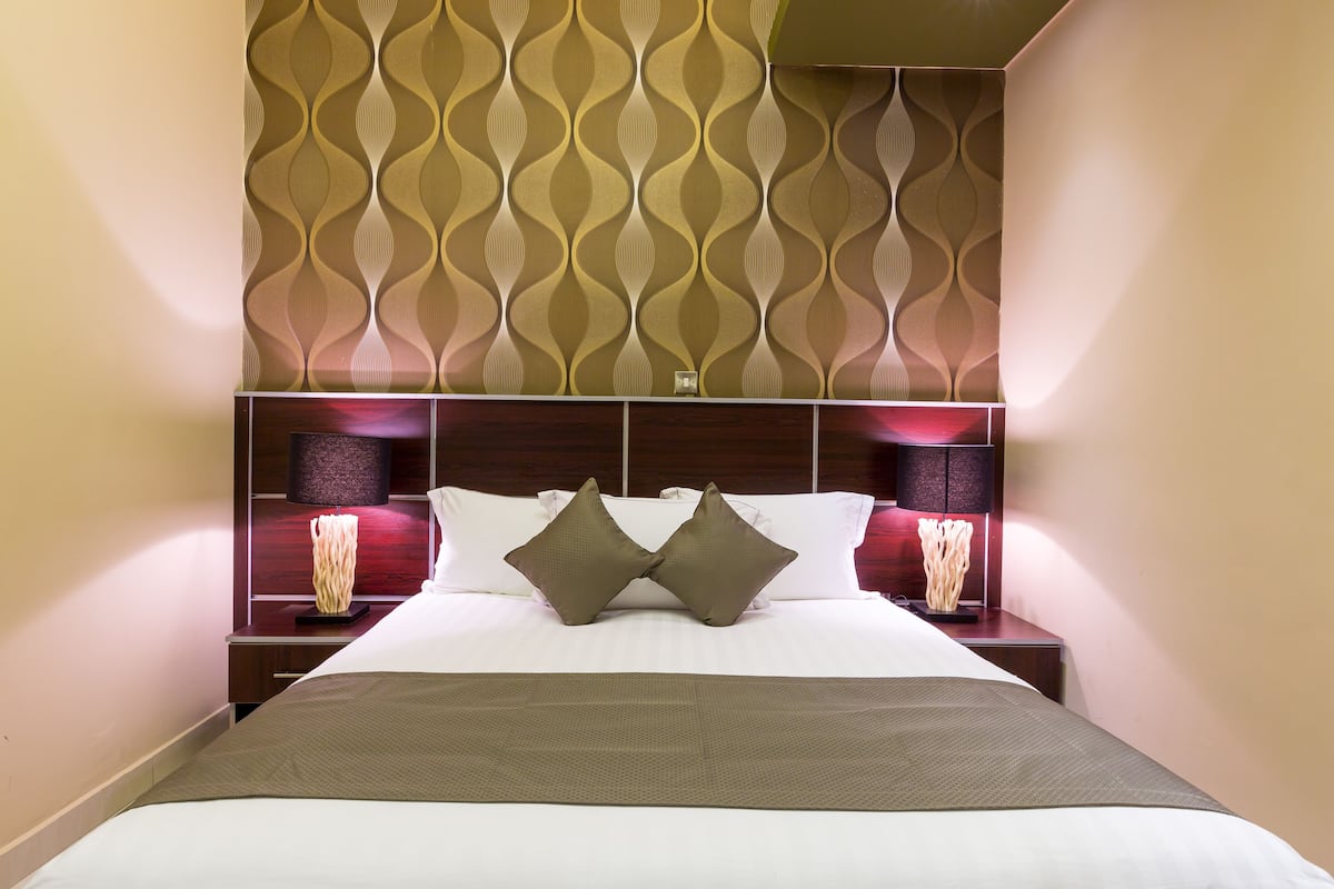 A hotel room with a patterned wallpaper, a neatly made bed with two pillows, and bedside lamps.