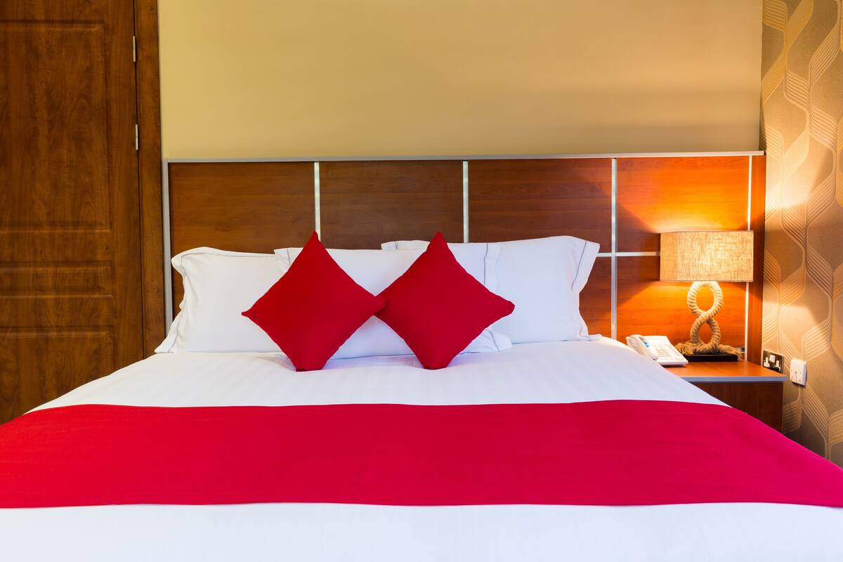 A neatly made bed with white and red bedding, a wooden headboard, and a bedside table with a lamp and phone.