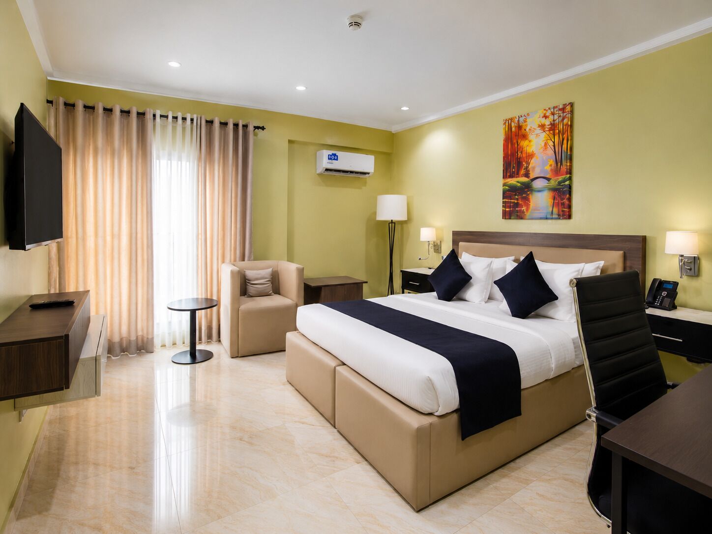Une chambre d’hôtel comprenant un lit, une télévision, un bureau, une chaise, une lampe, un appareil de climatisation et un tableau au mur.