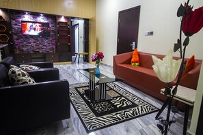 Una sala de estar moderna con un sofá, una mesa de centro y un televisor montado en una pared de ladrillo. Hay una puerta que conduce a otra habitación y un jarrón con flores sobre la mesa de centro.