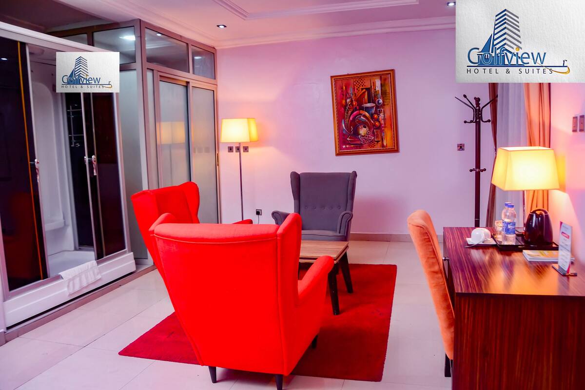 A hotel room with a red chair, a grey armchair, a wooden table, and a desk with a lamp. There is a painting on the wall and a coat rack.