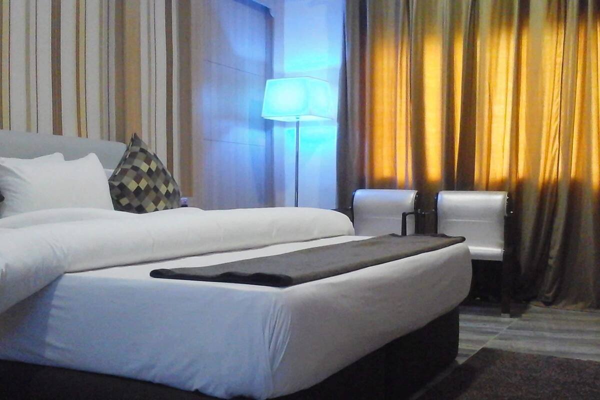 A hotel room with a large bed, pillows, a bedside table, a lamp, and curtains.