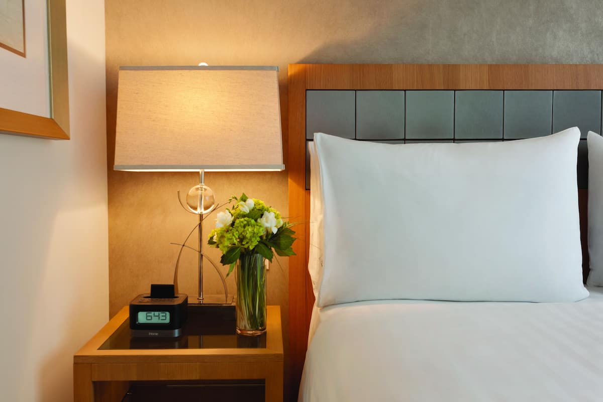 Une chambre d’hôtel avec un lit, une table de chevet avec une lampe, une horloge numérique et un vase avec des fleurs.