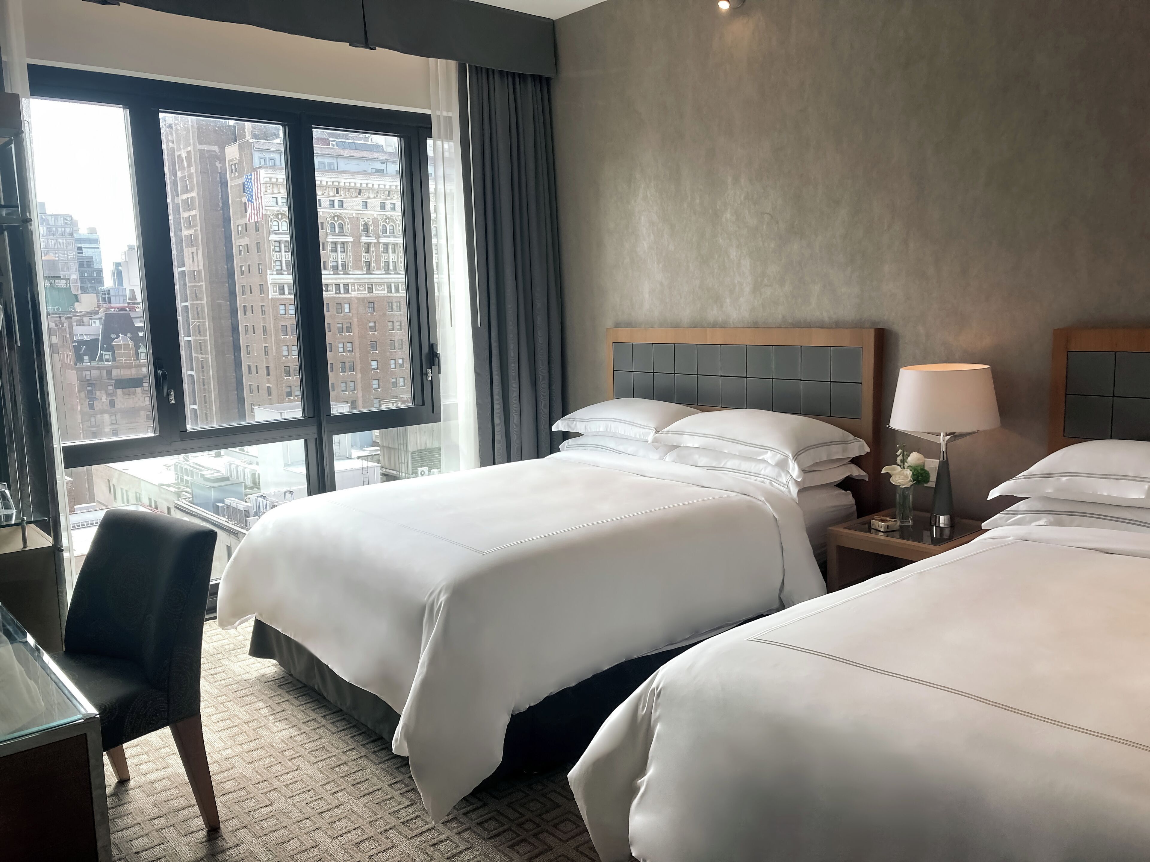 Habitación de hotel con dos camas, un escritorio y vistas a edificios de la ciudad.