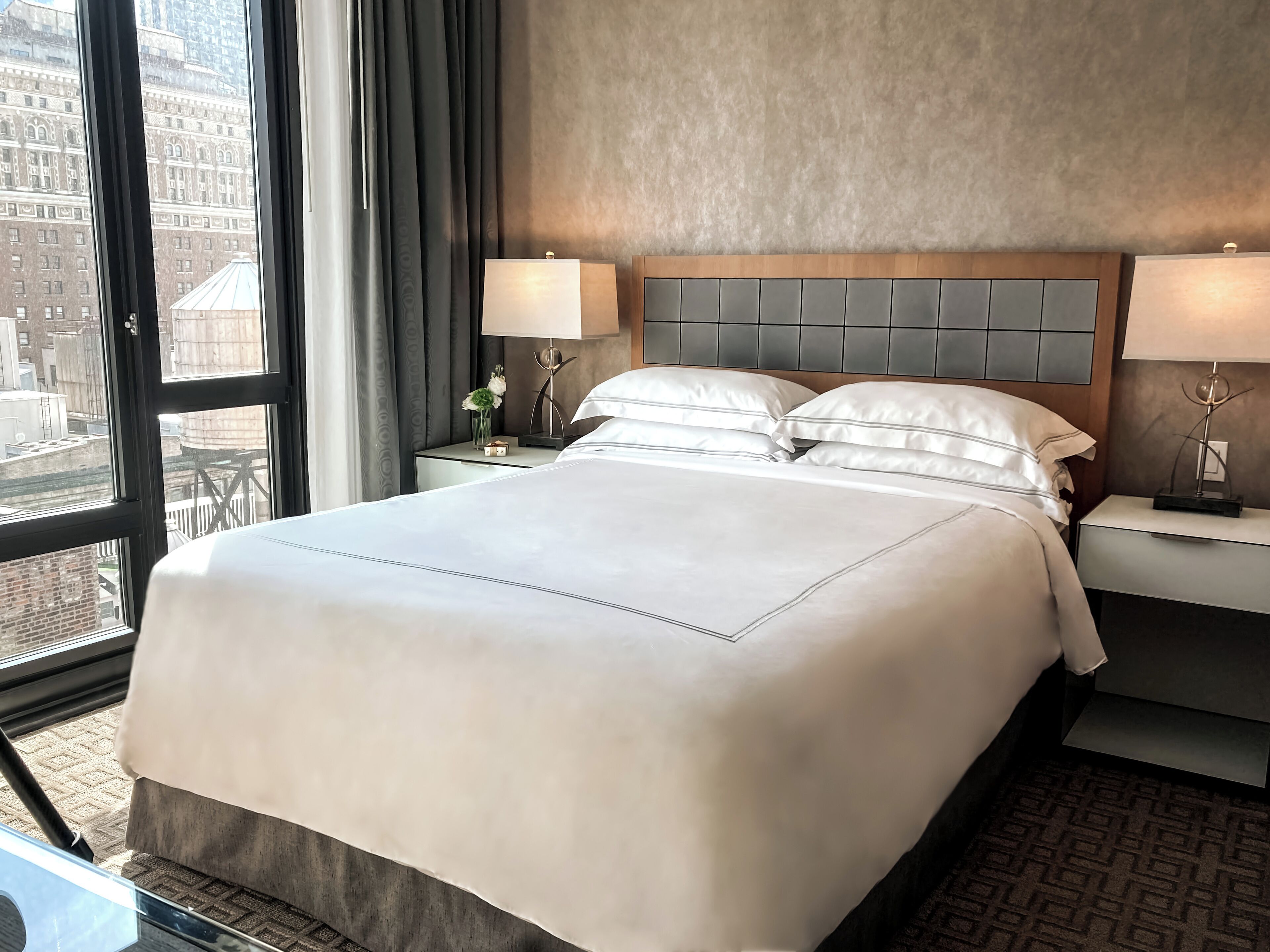 Habitación de hotel con una cama grande, dos mesitas de noche con lámparas, vista a la ciudad a través de amplias ventanas y una pared con textura.
