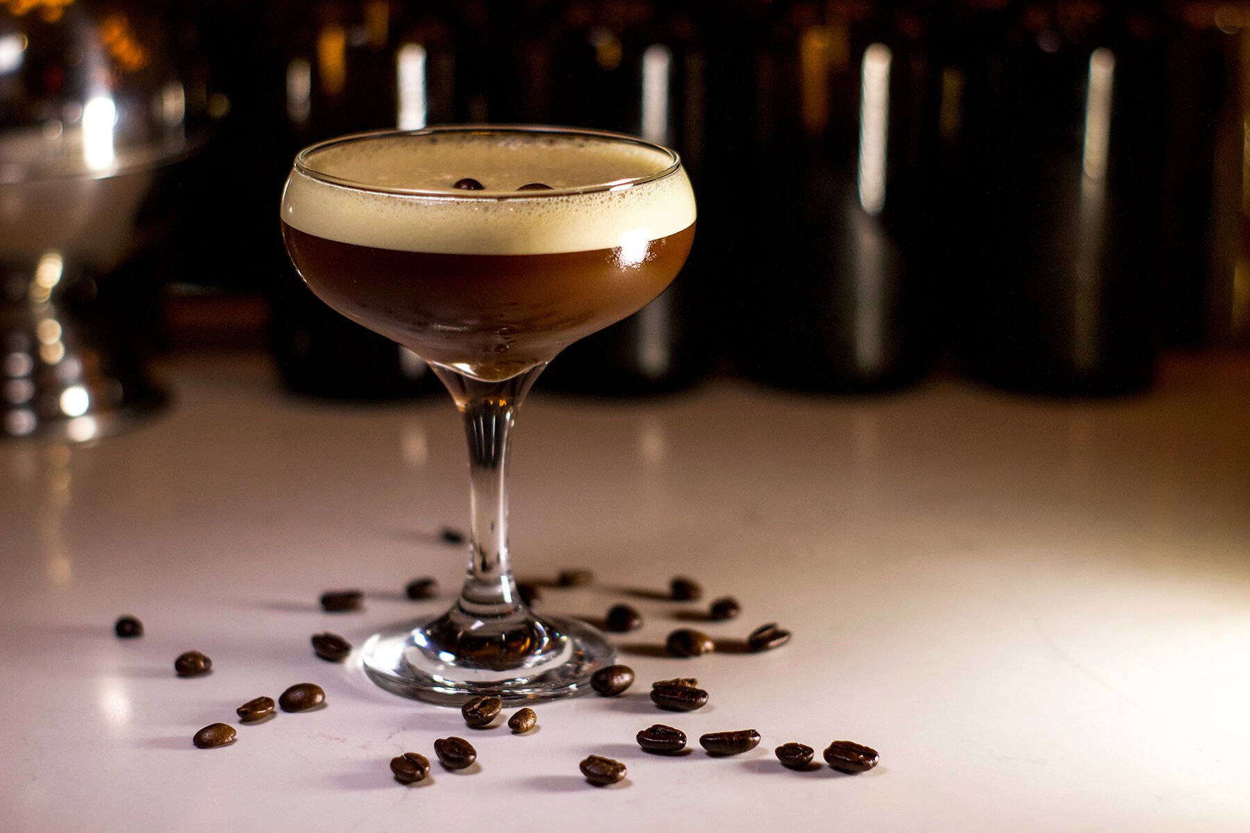 Una taza de líquido oscuro con espuma, rodeada de granos de café sobre una superficie blanca.