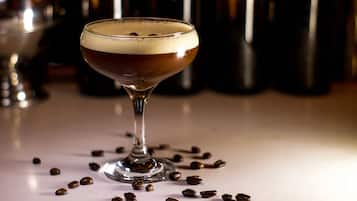 Una taza de líquido oscuro con espuma, rodeada de granos de café sobre una superficie blanca.