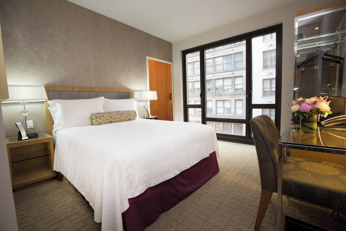 Ein Hotelzimmer mit Bett, Nachttischen, Schreibtisch mit Stuhl, Fenster mit Blick auf Gebäude und einer Tür.