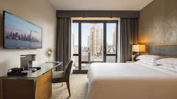 A hotel room with a large bed, a desk with a coffee maker, a chair, and a large window with a city view.