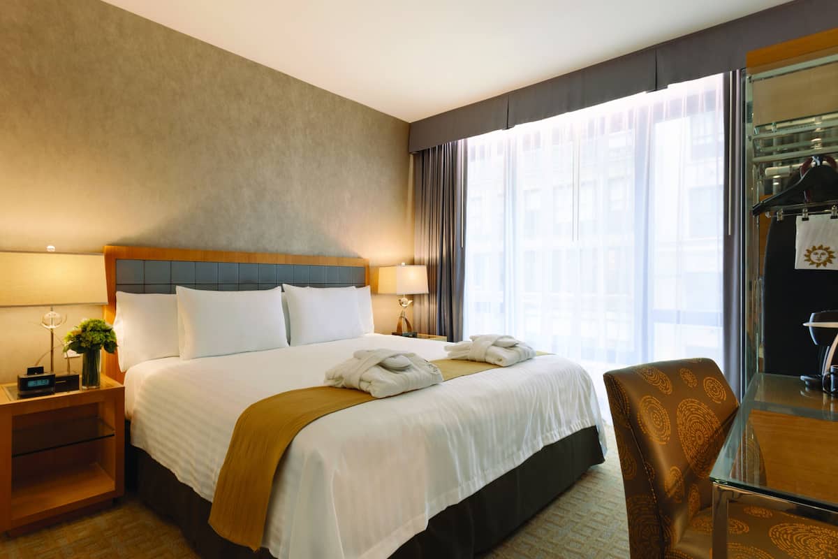 Une chambre d’hôtel avec un grand lit, une table de chevet avec une lampe, une chaise et une vue sur la ville grâce à de grandes fenêtres.