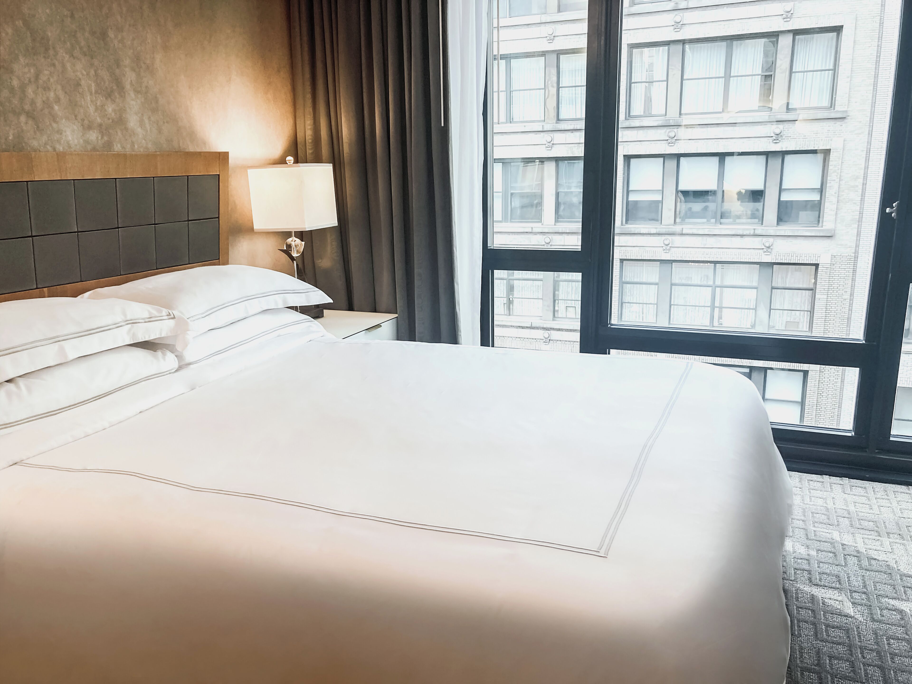 A hotel room with a large bed, a nightstand with a lamp, and a view of buildings through a large window.