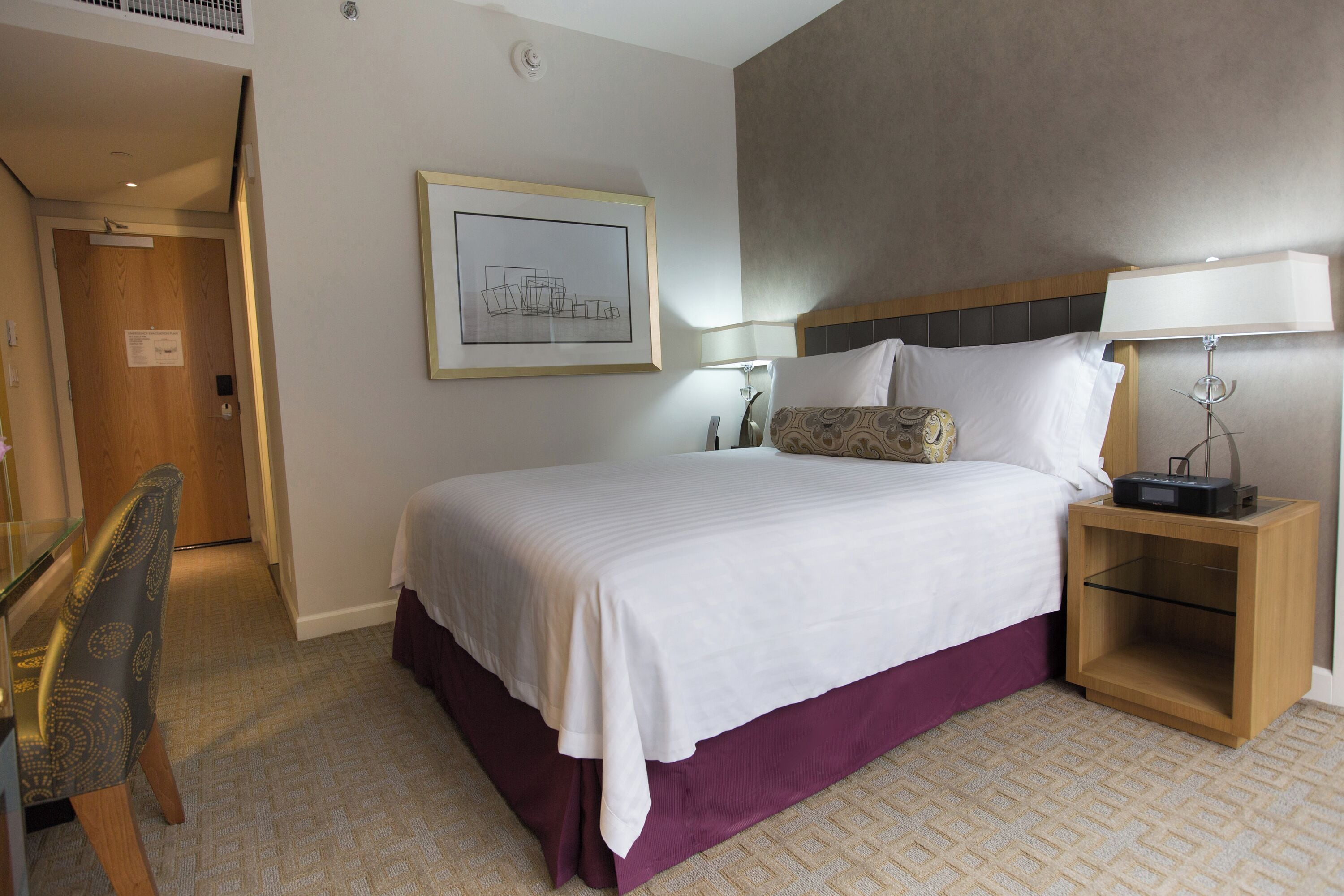 Une chambre d’hôtel avec un lit, une table de chevet, une lampe, une chaise et un tableau encadré au mur.