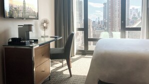 Een hotelkamer met een bed, een bureau met koffiezetapparaat, een stoel en uitzicht op de stad door grote ramen, plus een schilderij van een stadsgezicht.