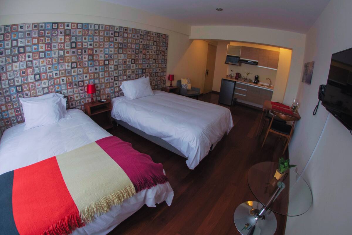 Quarto de hotel com duas camas, uma kitchenette pequena e uma televisão presa na parede.