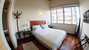 Un dormitorio con una cama grande, una mesita de noche, una lámpara, un espejo y una ventana con vistas a edificios.