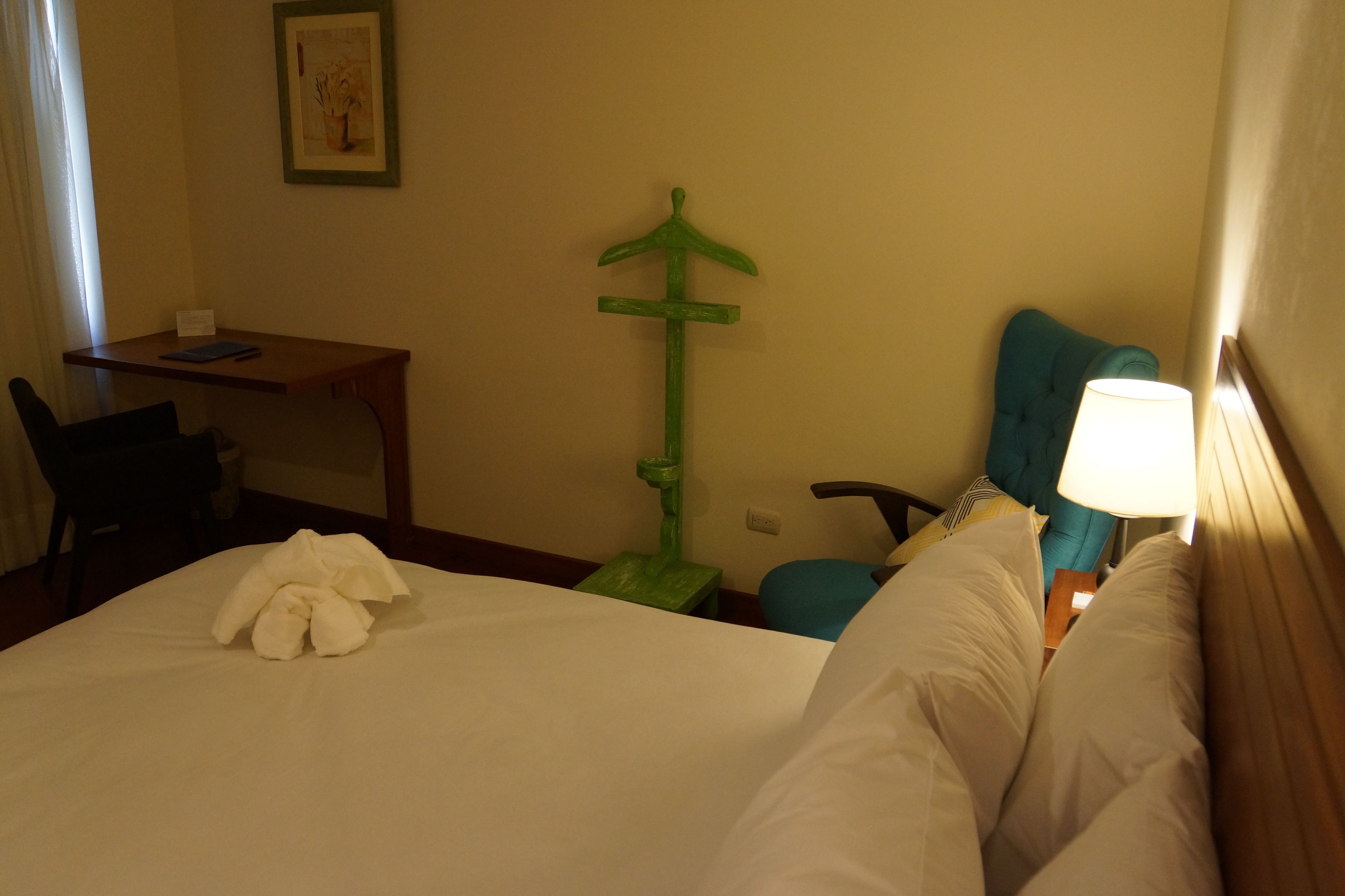 A hotel room with a bed, a desk, a chair, a green decorative item, and a lamp.