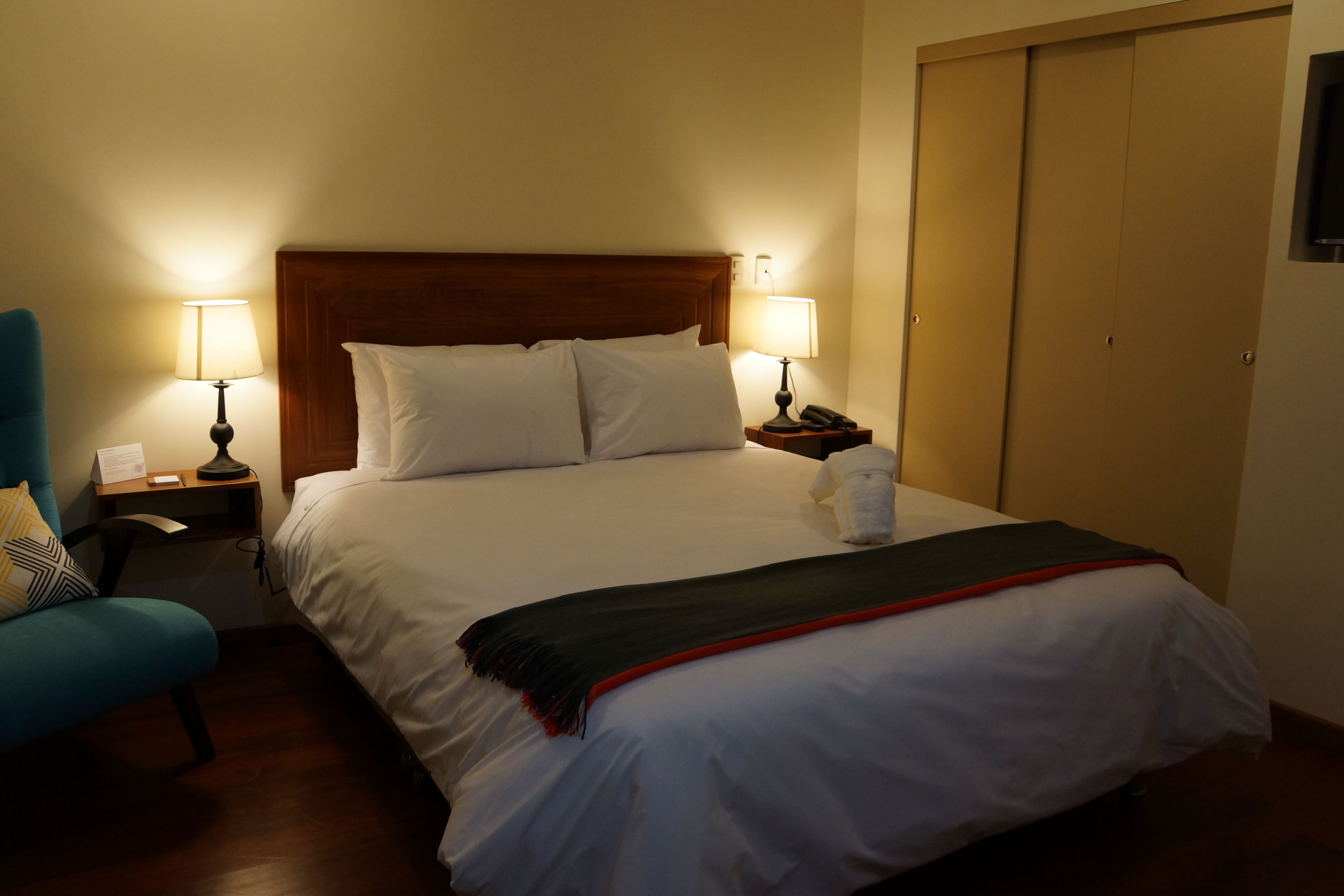 A neatly made bed with white linens and a red blanket, flanked by two bedside tables with lamps, a nightstand, and a blue chair.