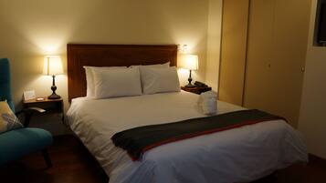 A neatly made bed with white linens and a red blanket, flanked by two bedside tables with lamps, a nightstand, and a blue chair.